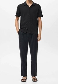 Chemise noire à manches courtes en tissu texturé, boutonnée sur le devant, associée à un pantalon noir ample et des sandales plates noires.