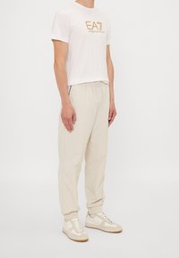 Herroutfit med en vit t-shirt med guldtryck, beige joggingbyxor med elastisk midja och ljusa sneakers med en texturerad design och gummisula.