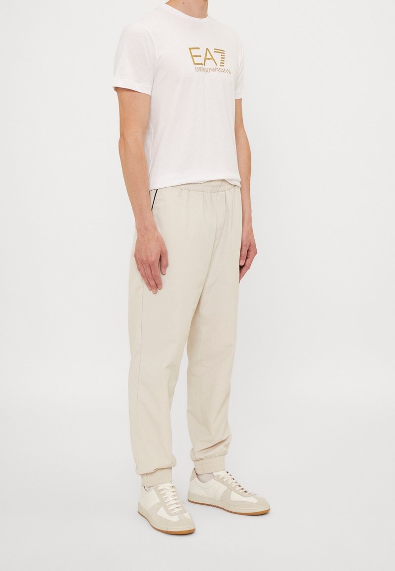 Herroutfit med en vit t-shirt med guldtryck, beige joggingbyxor med elastisk midja och ljusa sneakers med en texturerad design och gummisula.