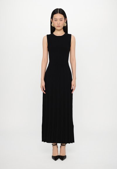 Robe maxi noire sans manches avec un ribbing vertical, corsage ajusté, jupe évasée et texture subtile. Une paire d'escarpins noirs à bout pointu.