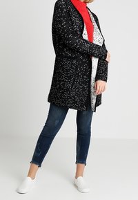 Czarny dziergany kardigan o strukturalnym wzorze, noszony na czerwonym golfie, w połączeniu z niebieskimi dżinsami i białymi butami typu slip-on.