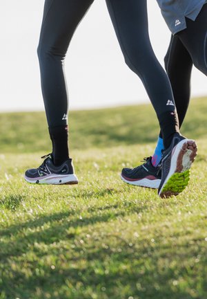 Schwarze Laufschuhe mit strukturierten Oberflächen und weißen Sohlen, getragen mit enganliegenden schwarzen Leggings und bunten Socken, auf grünem Terrain.