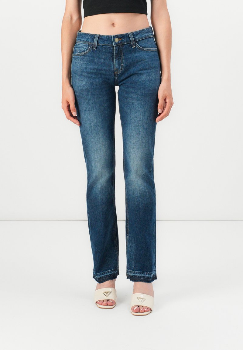 Egy nő közepes magasítóval rendelkező, egyenes lábú, enyhén kopott kék farmernadrágot visel, amit fehér, nyitott orrú magassarkú szandálokkal párosít, egy sima fehér háttér előtt.