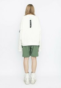 Sudadera oversized crema con etiqueta negra en la parte trasera, combinada con pantalones cortos verdes y botas blancas de plataforma hasta el tobillo. Textura de tela suave.