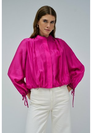 Donna che indossa una vivace blusa fucsia con maniche lunghe e a palloncino e pantaloni bianchi, in piedi davanti a uno sfondo grigio chiaro uniforme.