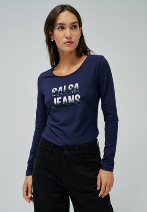 Camisa de manga larga azul marino con un cuello redondo, que presenta un brillo en la impresión de "Salsa Jeans" en blanco y azul en el pecho, combinada con pantalones negros.