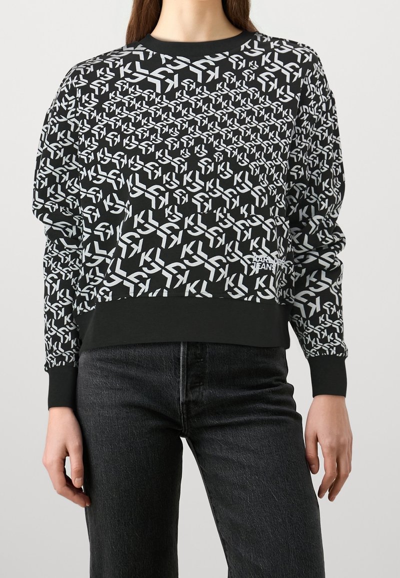 Mujer que lleva una sudadera negra con un patrón geométrico blanco de "KL" y el texto "KARL JEANS", combinada con jeans oscuros.