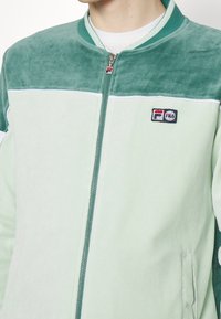 Zielona welurowa kurtka na zamek z dopasowanym kołnierzem, kontrastowymi białymi paskami i logo na piersi. Miękka w dotyku i o luźnym kroju.