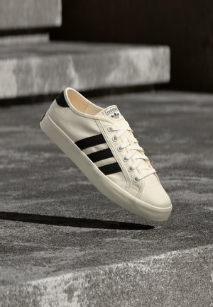 Λευκό αθλητικό Adidas με μαύρες ρίγες φαίνεται να αιωρείται πάνω από τσιμεντένια σκαλιά, τοποθετημένο με τρόπο που φαίνεται η πλευρά και τα κορδόνια στο φως του ήλιου.
