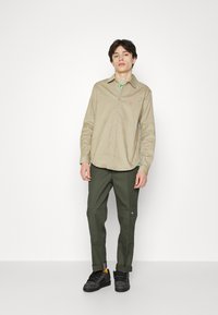 Camisa de botón color marrón claro con cuello, combinada con pantalones oscuros verdes. La camisa tiene un logotipo bordado en el lado izquierdo del pecho. Zapatillas negras.