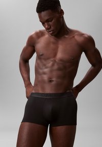 Czarne bawełniane bokserki o gładkiej strukturze z paskiem w logo Calvin Klein. Model przyjmuje neutralną pozę, na szarym tle.