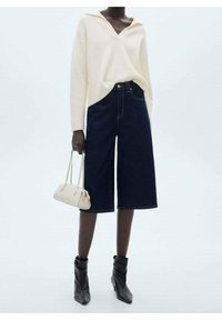 Übergroßer cremefarbener Strickpullover mit V-Ausschnitt, kombiniert mit dunklen Denim-Culottes. Die Accessoires umfassen eine kleine hellbeige Handtasche und schwarze Stiefeletten.