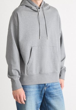 Person iført en ensfarvet grå hættetrøje med forlomme og lyseblå jeans, stående mod en hvid baggrund.