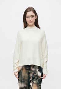 Kremowy, lekki sweter dzianinowy z wysokim kołnierzem i oversize'owymi rękawami, zestawiony z luźnymi spodniami w wzory.