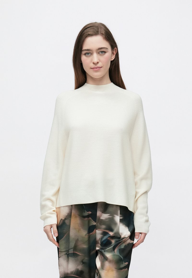 Kremowy, lekki sweter dzianinowy z wysokim kołnierzem i oversize'owymi rękawami, zestawiony z luźnymi spodniami w wzory.