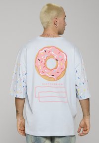 Koszulka z krótkim rękawem w jasnoniebieskim kolorze, z du�żą różową grafiką pączka na plecach, posypaną kolorowymi posypkami; luźny krój i miękka faktura.