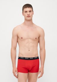 Röda bomullsnättpants med svart elastisk midja som har varumärket "HUGO" i vitt. Slät textur, åtsittande design.