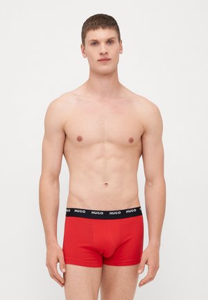Rote Baumwoll-Boxershorts mit einem schwarzen Elastikbund, auf dem der Markenname "HUGO" in Weiß dargestellt ist. Glatte Textur, eng anliegendes Design.