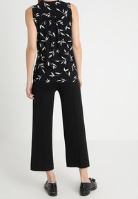 Top negro sin mangas con estampado de hojas blancas, con una parte trasera fruncida; combinado con pantalones anchos negros y zapatos negros.