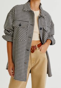 Houndstooth geruite overhemdjas in zwart en wit, met een kraag, knoopsluiting aan de voorkant en borstzakken. Draagt beige broek eronder.