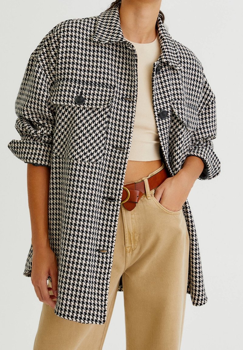 Houndstooth geruite overhemdjas in zwart en wit, met een kraag, knoopsluiting aan de voorkant en borstzakken. Draagt beige broek eronder.