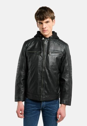 Junger Mann trägt eine schwarze Lederjacke mit Reißverschluss vorne, Reißverschlusstaschen auf der Brust und einer schwarzen Kapuze, kombiniert mit blauen Jeans.