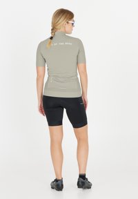 Vaaleahiuksinen nainen, jolla on plaitattu letti, pukeutunut harmaaseen lyhythihaiseen pyöräilypaitaan ja mustiin pehmustettuihin shortseihin, seisoo selin valkoista taustaa vasten.