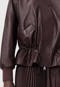 Burgundská kožená bunda s žebrovanými detaily, elastickým pasem a bočním kapsou. Přiléhavé manžety a měkký, texturovaný povrch.