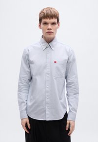 Φόρεμα με κουμπιά σε ανοιχτό γαλάζιο χρώμα, με μυτερό γιακά, μακριά μανίκια, τσέπη στο στήθος και κόκκινη ετικέτα μάρκας. Λείο ύφασμα, στενή εφαρμογή, μίνιμαλ σχεδίαση.