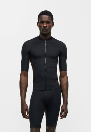 Homme debout face à l'objectif portant un maillot de cyclisme noir ajusté à manches courtes et un short cycliste noir assorti sur un fond uni.