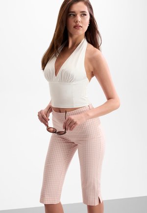 Junge Frau in weißem Neckholdertop und rosa karierten Caprihosen hält Sonnenbrille und schaut seitlich vor schlichtem Hintergrund.