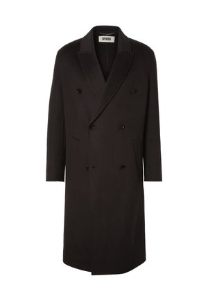 Jeune homme debout portant un long manteau croisé marron foncé, une chemise beige, un pantalon en velours côtelé marron foncé et des bottes en cuir noir.