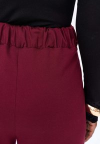 Bordeauxkleurige broek met een elastische tailleband; gladde stofstructuur; naad die verticaal aan de achterkant loopt; gecombineerd met een zwarte top.