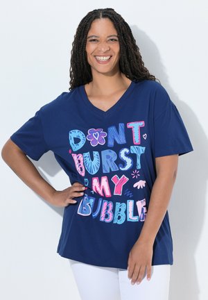 Lachende vrouw met gevlochten haar draagt een blauw shirt met de tekst "Don't burst my bubble" in kleurrijke, speelse letters, poserend met één hand op de heup.