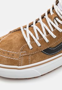 Tenisi din suede maro cu șireturi și cusături albe. Prezintă un logo în formă de swoosh negru de-a lungul lateralului și o talpă din cauciuc albă cu textură pentru aderență.