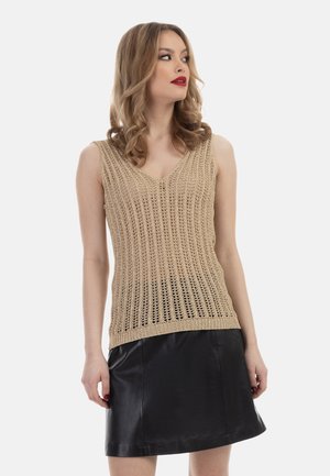 Kvinde med krøllet hår iført en beige strikket ærmeløs top og sort lædernederdel, kigger mod højre mod en hvid baggrund.