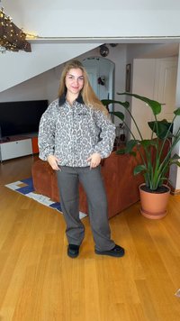 Harmaa leopardi-kuvioitu vetoketjullinen takki mustalla kauluksella, yhdistettynä harmaisiin housuihin ja mustiin kenkiin, asetettuna kasvien ja sohvan ympäröimään huoneeseen.