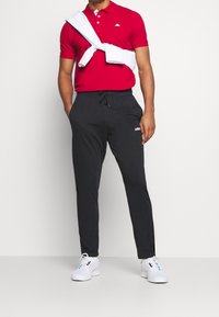 Polo rouge à manches courtes, pantalon de jogging noir et baskets blanches. Serviette posée sur les épaules. Logo présent sur le polo et le pantalon.