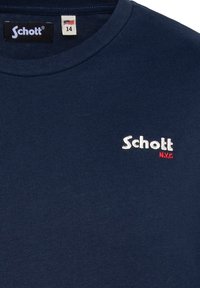 Camiseta de algodón azul marino con cuello redondo. Presenta el logo blanco "Schott" con acentos rojos. La etiqueta muestra el tamaño 14 y diseño de la bandera de EE. UU.
