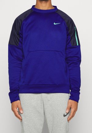 Homme portant un sweat-shirt bleu Nike avec des panneaux noirs aux épaules et un petit logo turquoise, associé à un pantalon de survêtement Nike gris clair.