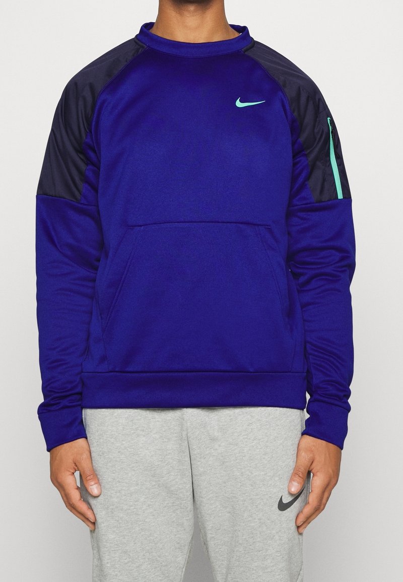 Homme portant un sweat-shirt bleu Nike avec des panneaux noirs aux épaules et un petit logo turquoise, associé à un pantalon de survêtement Nike gris clair.
