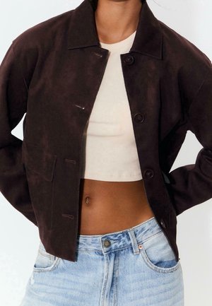 Persoon draagt een donkerbruine suède jas over een wit cropped rib topje en lichtblauwe high-waisted jeans.