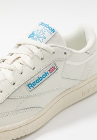 Baskets blanches Reebok Classic avec logo bleu, détail du drapeau Union Jack, lacets blancs et semelle texturée sur fond blanc uni.