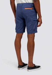 Pantaloni corti blu navy con una tasca posteriore dotata di una zip multicolore, realizzati in un tessuto leggero, lunghezza al ginocchio, indossati con sneaker nere.
