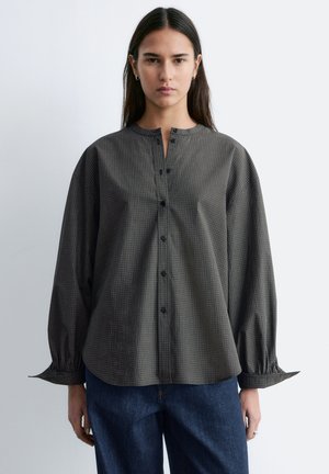 Jonge vrouw met lang donker haar draagt een losse, lange mouw, grijs geruite blouse met knopen en een blauwe spijkerbroek, staand tegen een effen achtergrond.