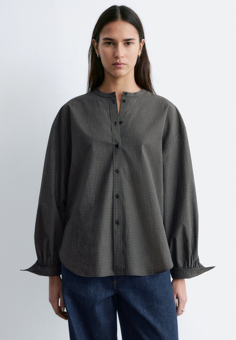 Jeune femme aux longs cheveux foncés portant une blouse ample à manches longues, à carreaux gris et boutonnée, ainsi qu'un jean bleu, debout devant un fond uni.