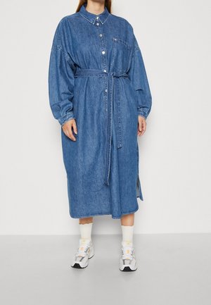 Person trägt ein langärmeliges, geknöpftes Jeanskleid mit Taillengürtel, weiße Socken und weiße Sneakers, steht vor einem schlichten Hintergrund.