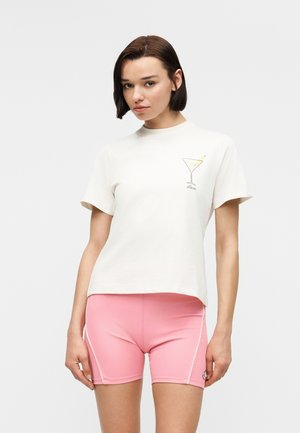 Femeie cu păr scurt și castaniu, purtând un tricou alb cu un model de pahar de cocktail și pantaloni scurți roz mulati, stând pe un fundal alb.