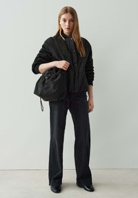 Giovane donna che indossa una giacca nera trapuntata e jeans neri a gamba larga, con un grande sacchetto nero a coulisse appeso a una spalla.