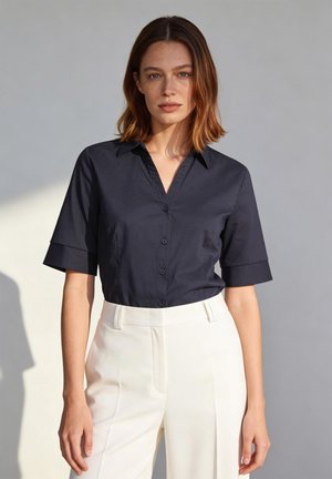 Femme aux cheveux bruns mi-longs portant une chemise noire à manches courtes avec des boutons et un pantalon blanc taille haute, debout devant un fond gris uni.